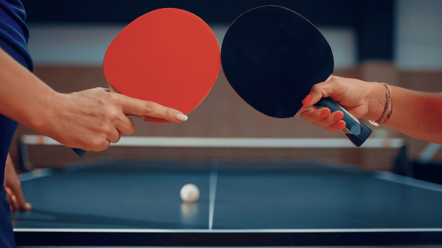 image of two table tennis rackets held up by two different hands with a table tennis ball sitting on a table tennis table in the background